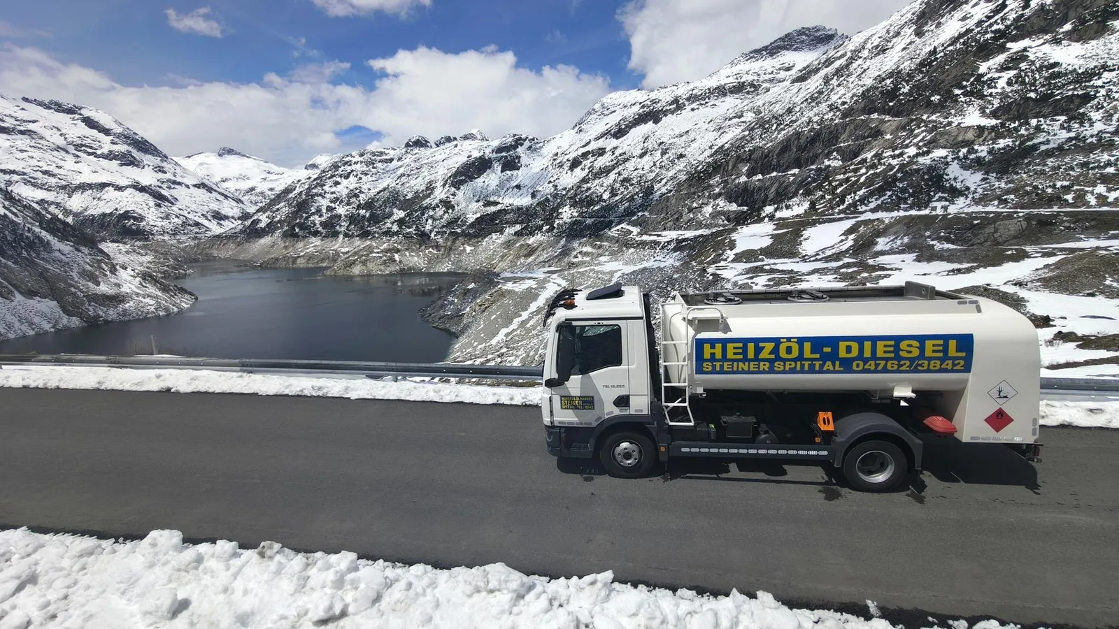 Tankwagen auf einer Bergstraße über einem Stausee, die Hänge noch mit Schnee