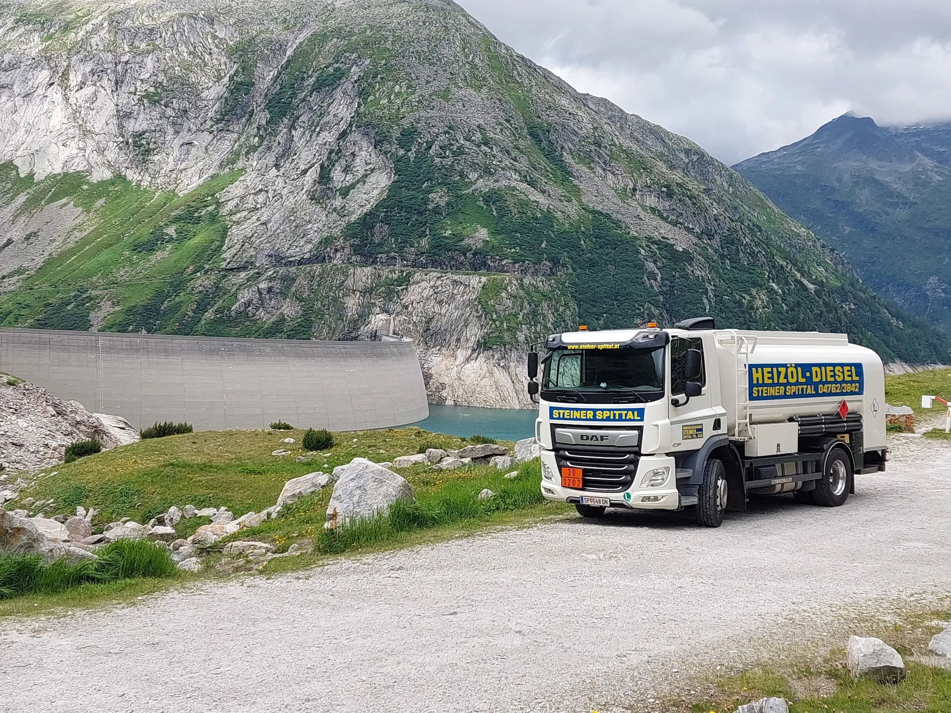 Weißer Tankwagen mit der Aufschrift Steiner Spittal, Heizöl-Diesel, auf einer Schotterstraße vor einer Staumauer und einem Bergsee